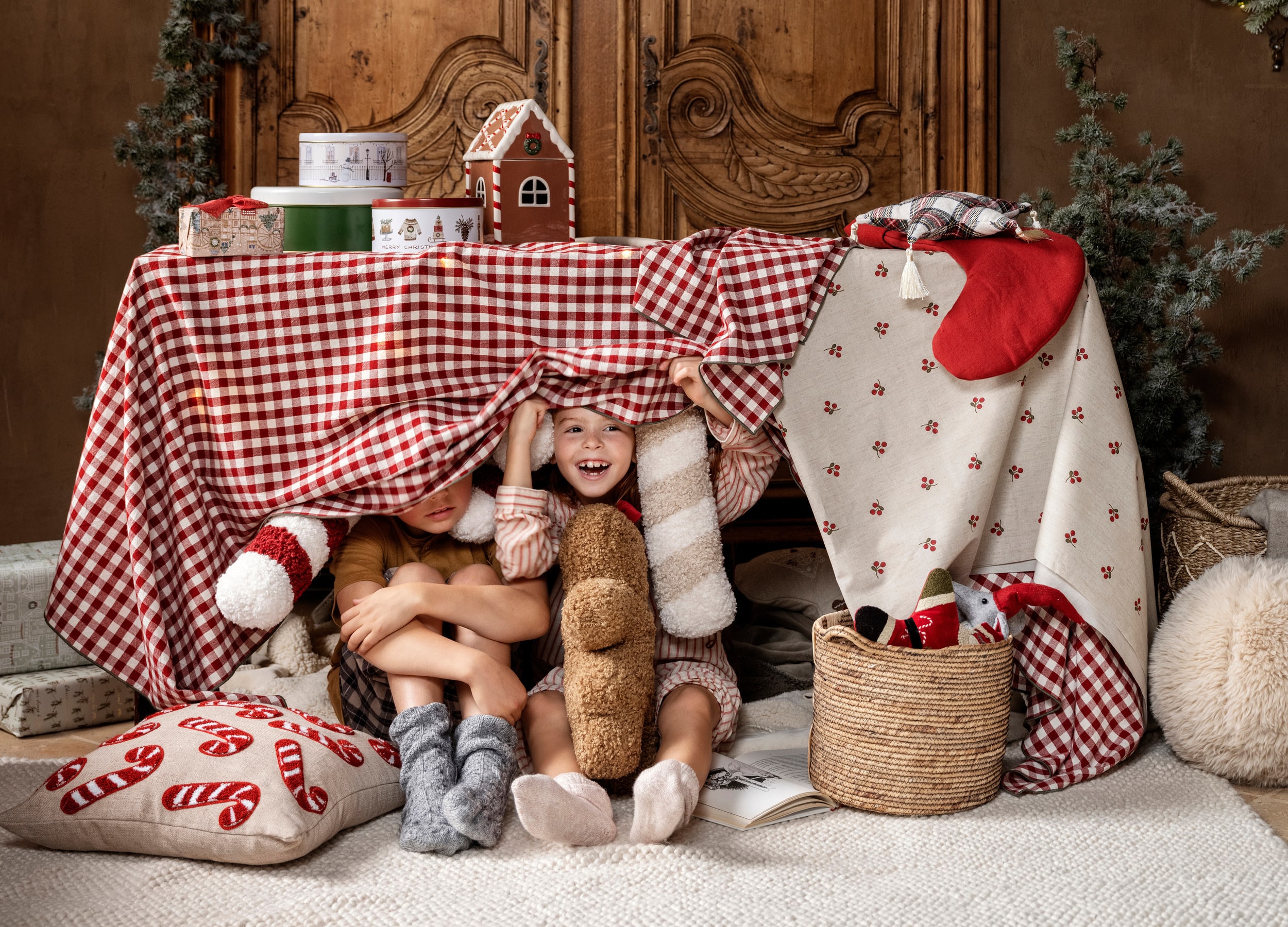 To barn sitter under et julepynta bord med duker, puter, bamser og diverse som hører jula til. Barna smiler.