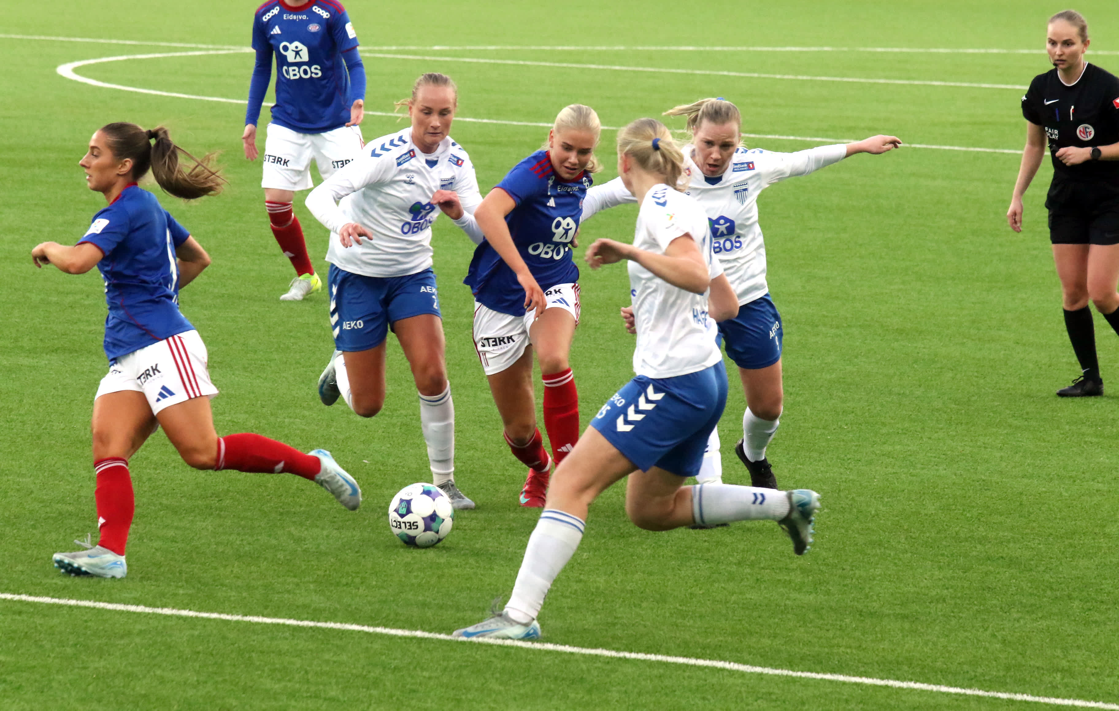 Foto av flere kvinnelige fotballspillere fra to lag som duellerer om ballen på banen.