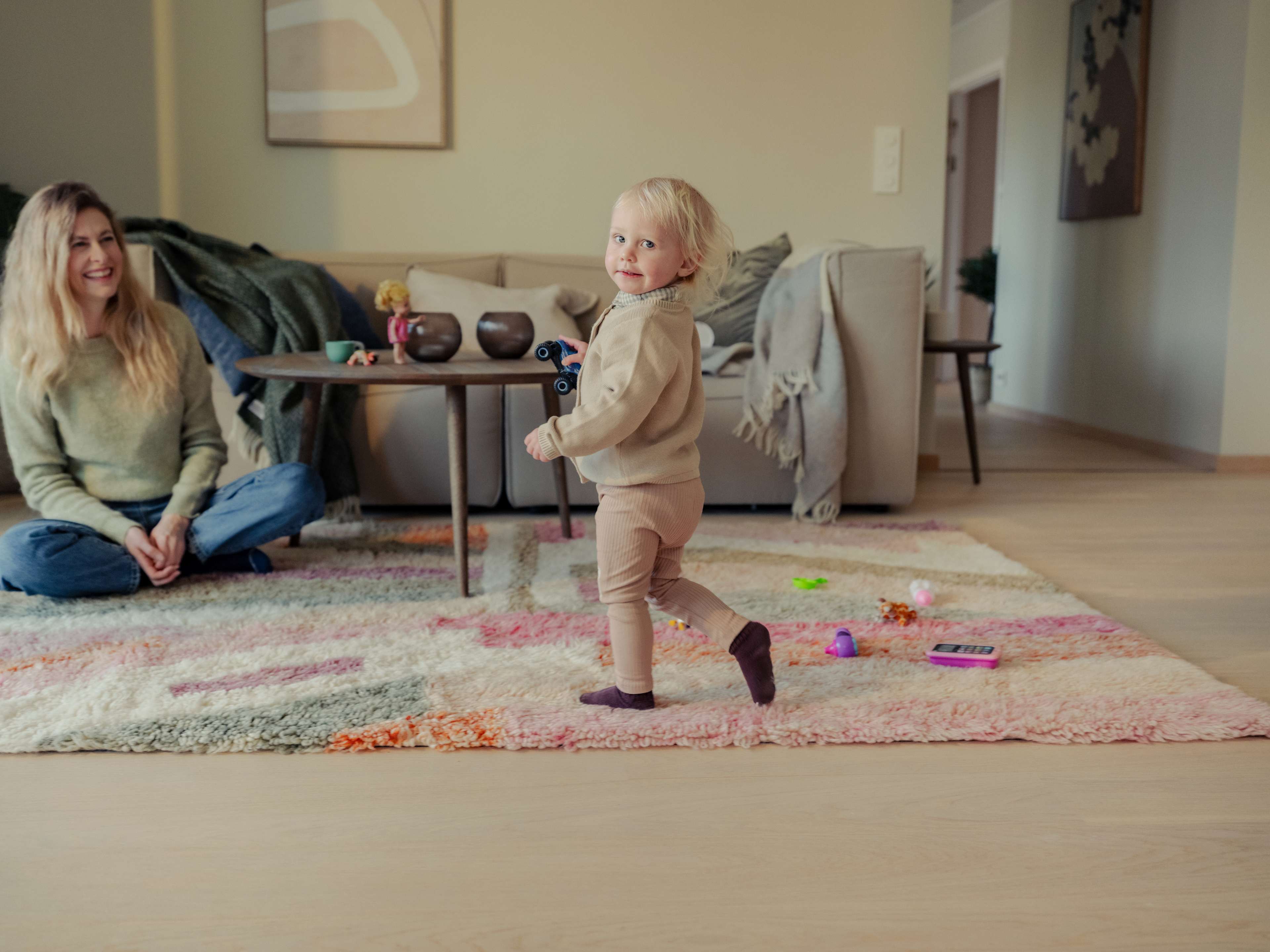 Jente som løper over et teppe og snur seg. Moren sitter på gulvet og ser på henne og smiler.