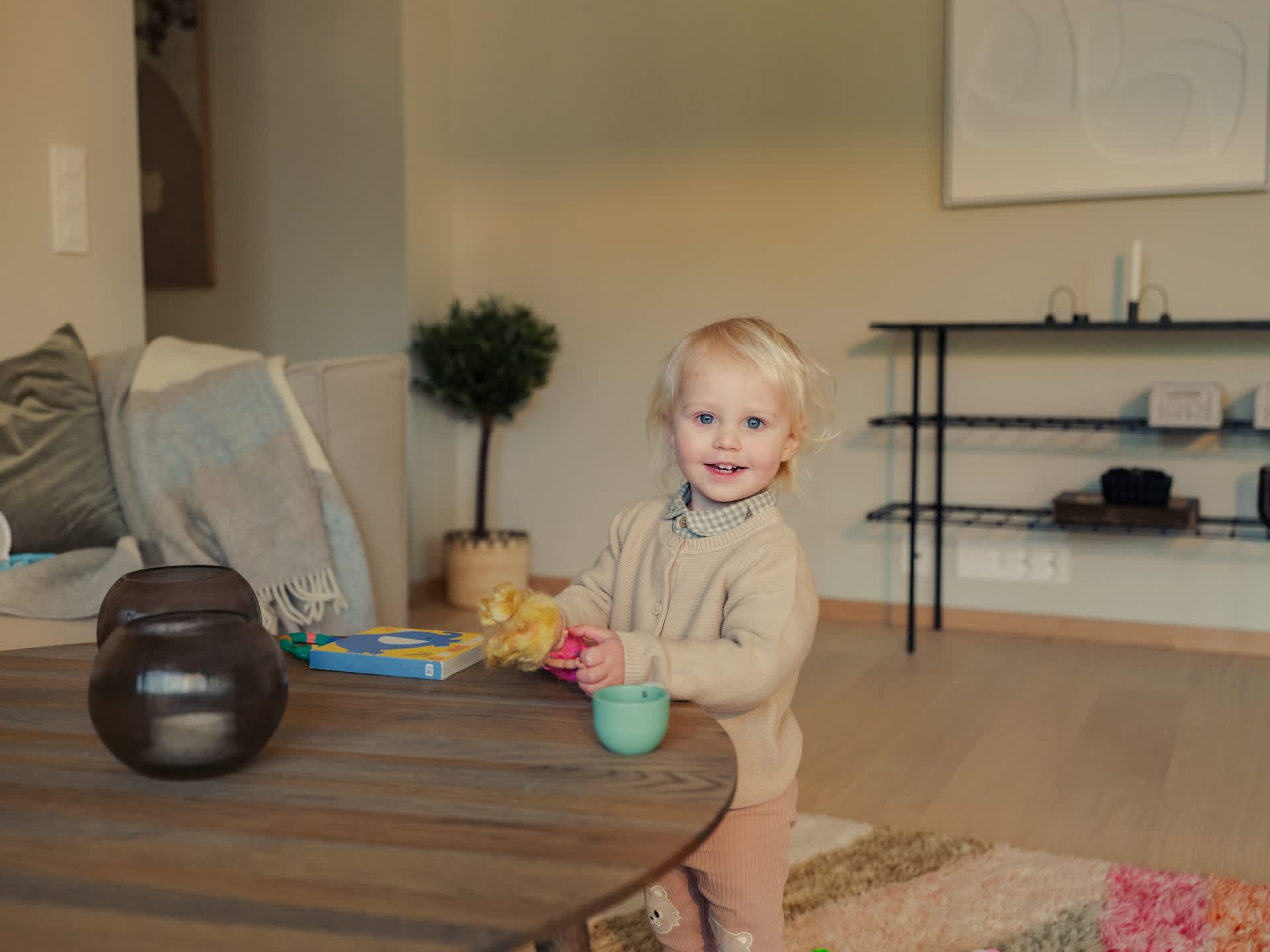 En jente har på seg beige genser og rosa bukse. Hun smiler. Hun er i en leilighet.
