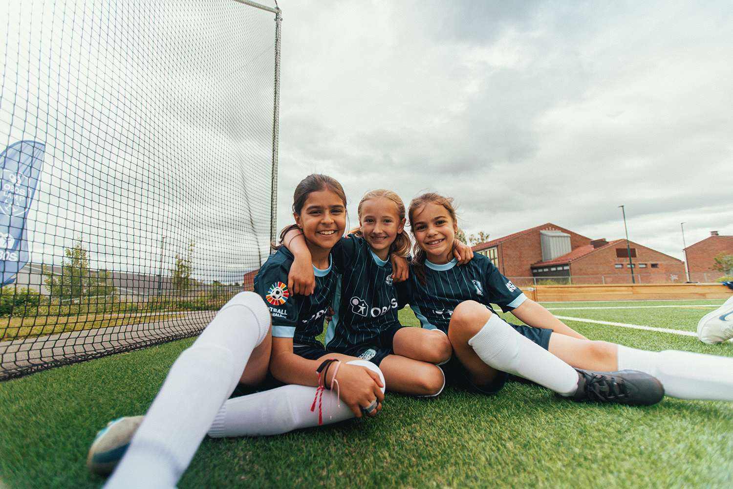 Tre jenter i fotballdrakter sitter ved siden av målet og smiler til kamera.