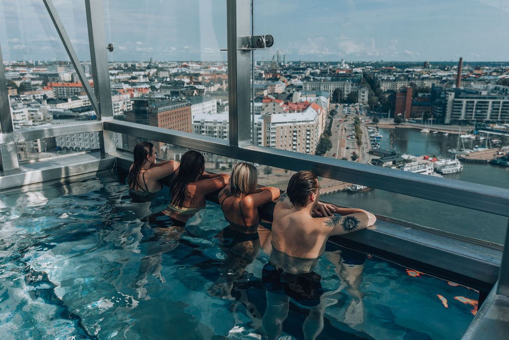 Fire personer bader i et basseng. De lener seg mot en kant og ser ut av vinduet utover byen.