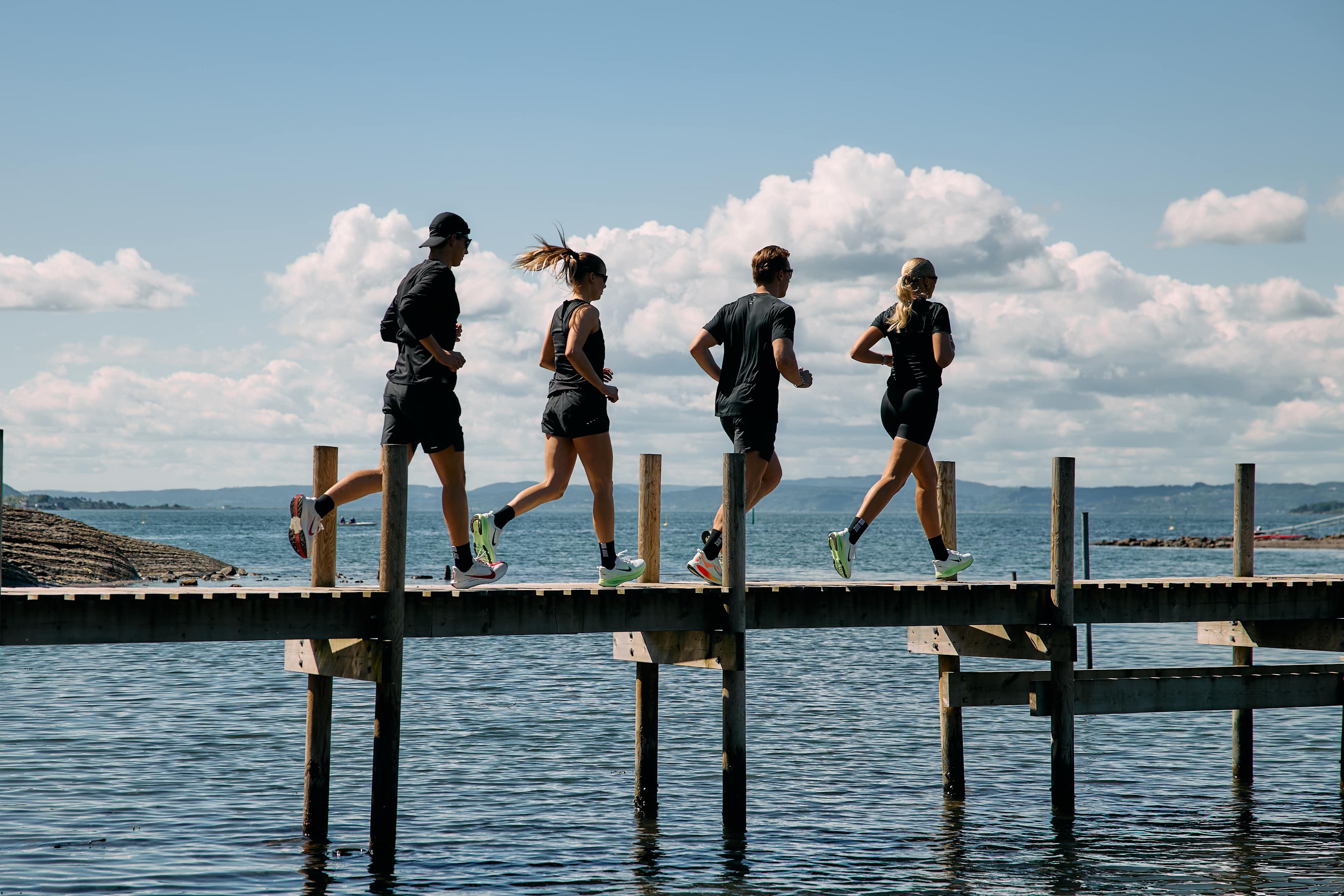 Fire personer løper på en brygge. De har alle på seg svarte treningsklær. Du ser fjorden ved siden av.