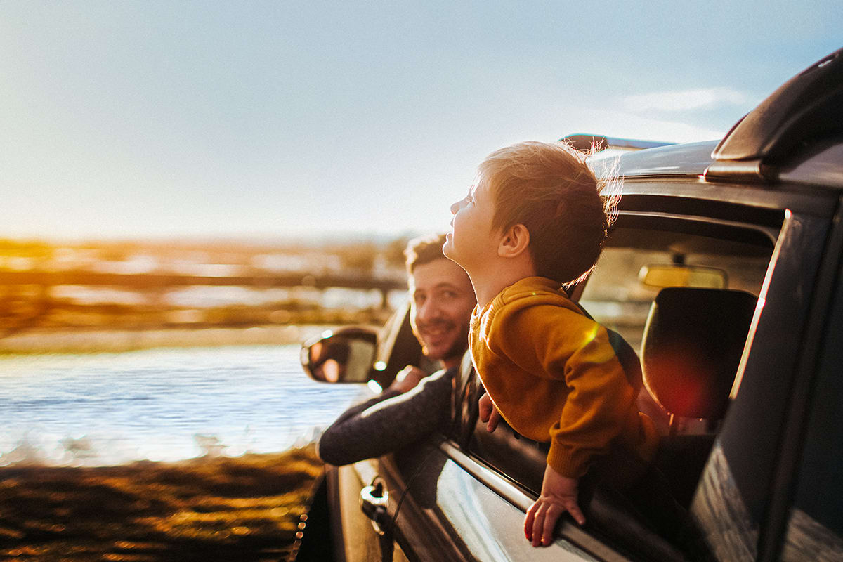 En gtt henger ut av vinduet på bilen og smiler. Faren snur seg og smiler også.