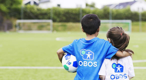En gutt og en jente med ryggen til står på en fotballbane. Gutten holder rundt jenta med høyre arm og har en fotball under venstre arm.