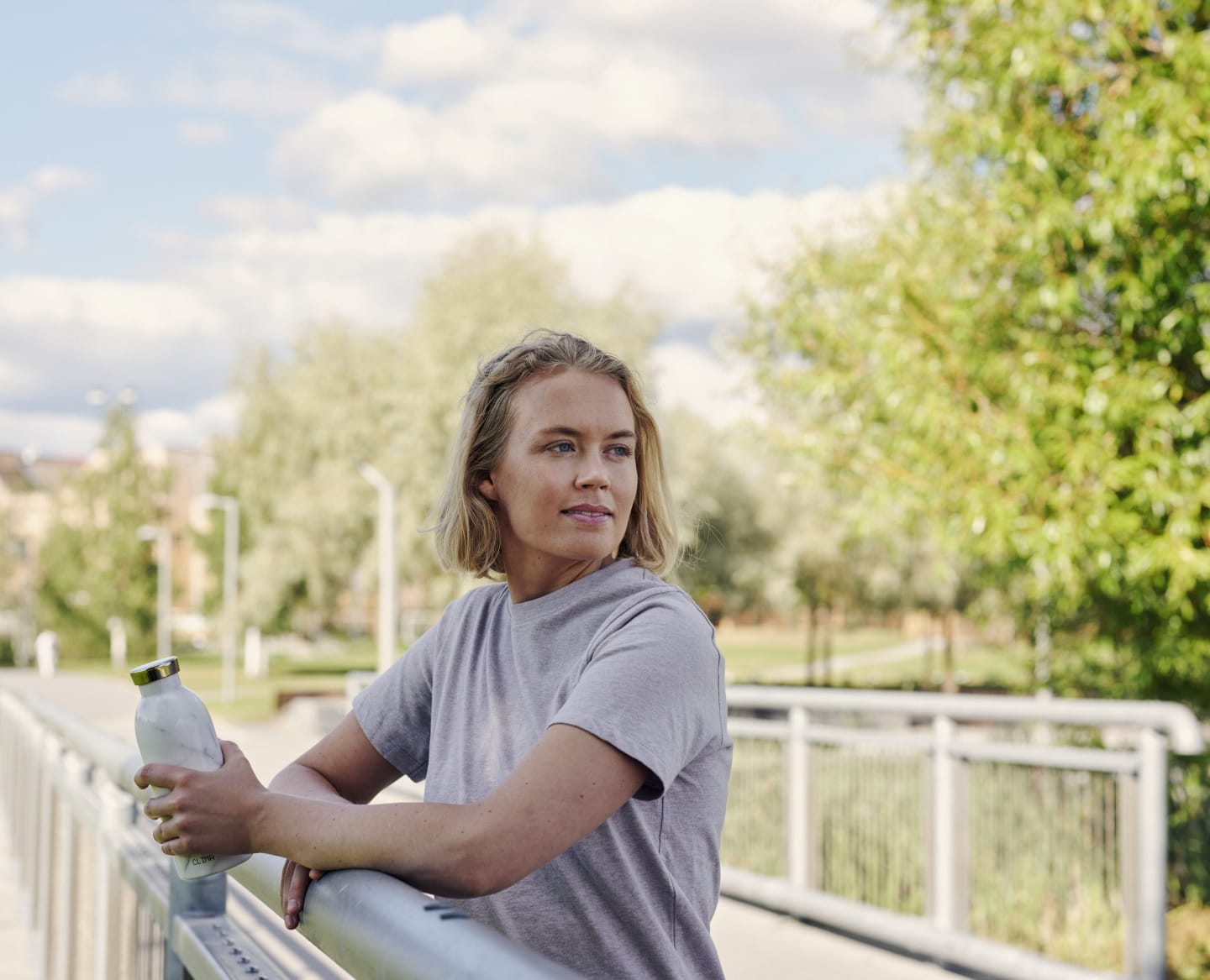 Ung kvinne holder en vannflaske i hånde og hviler seg mot rekkverket i en park.