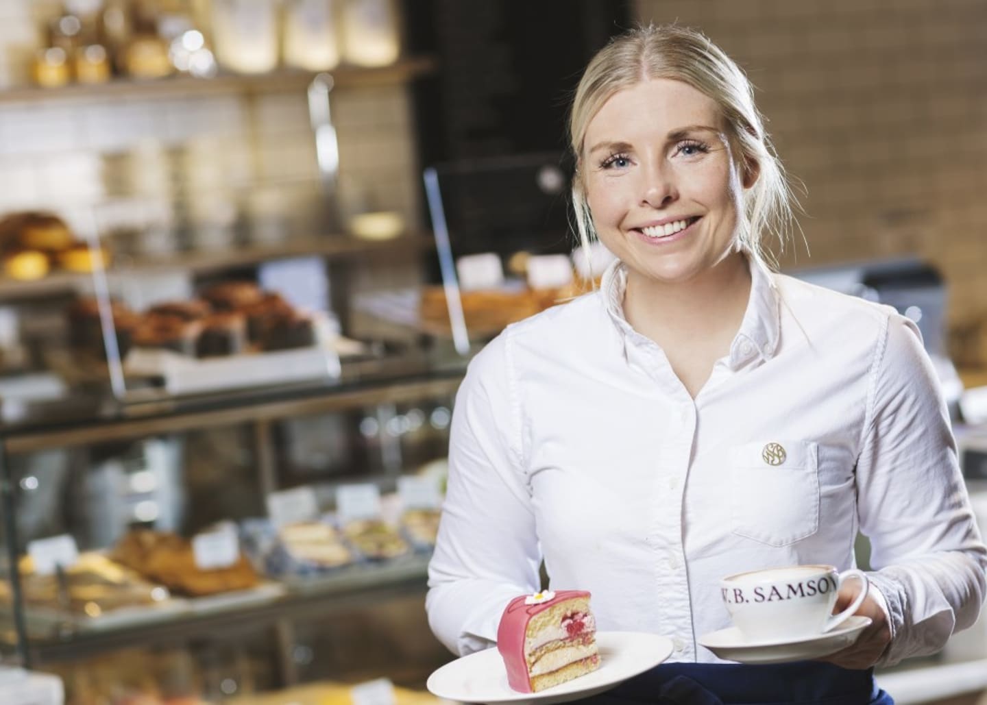 En servitør holder en asjett med kake og en kopp. Foto.