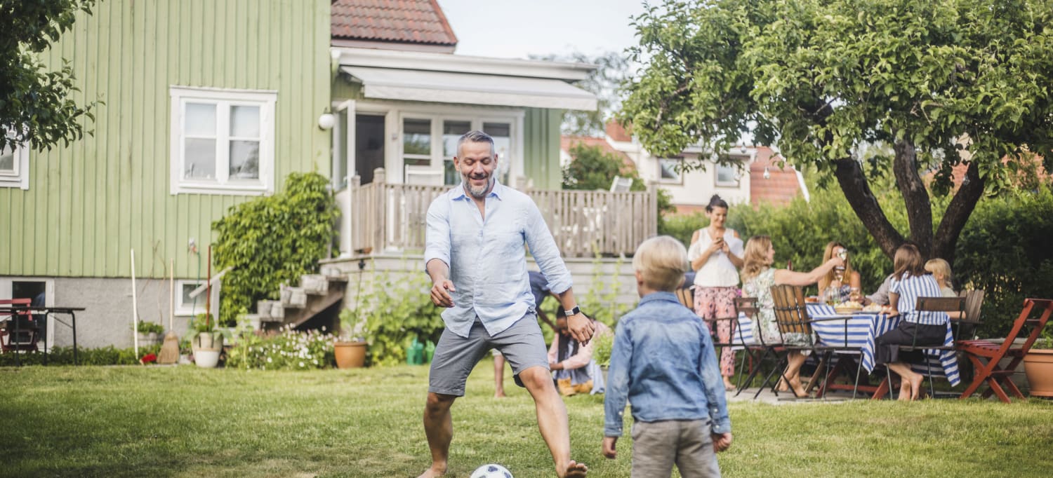 Foto av familie samlet i hage, med far og sønn som spiller fotball i forgrunnen, og god stemning ved bordet i bakgrunnen