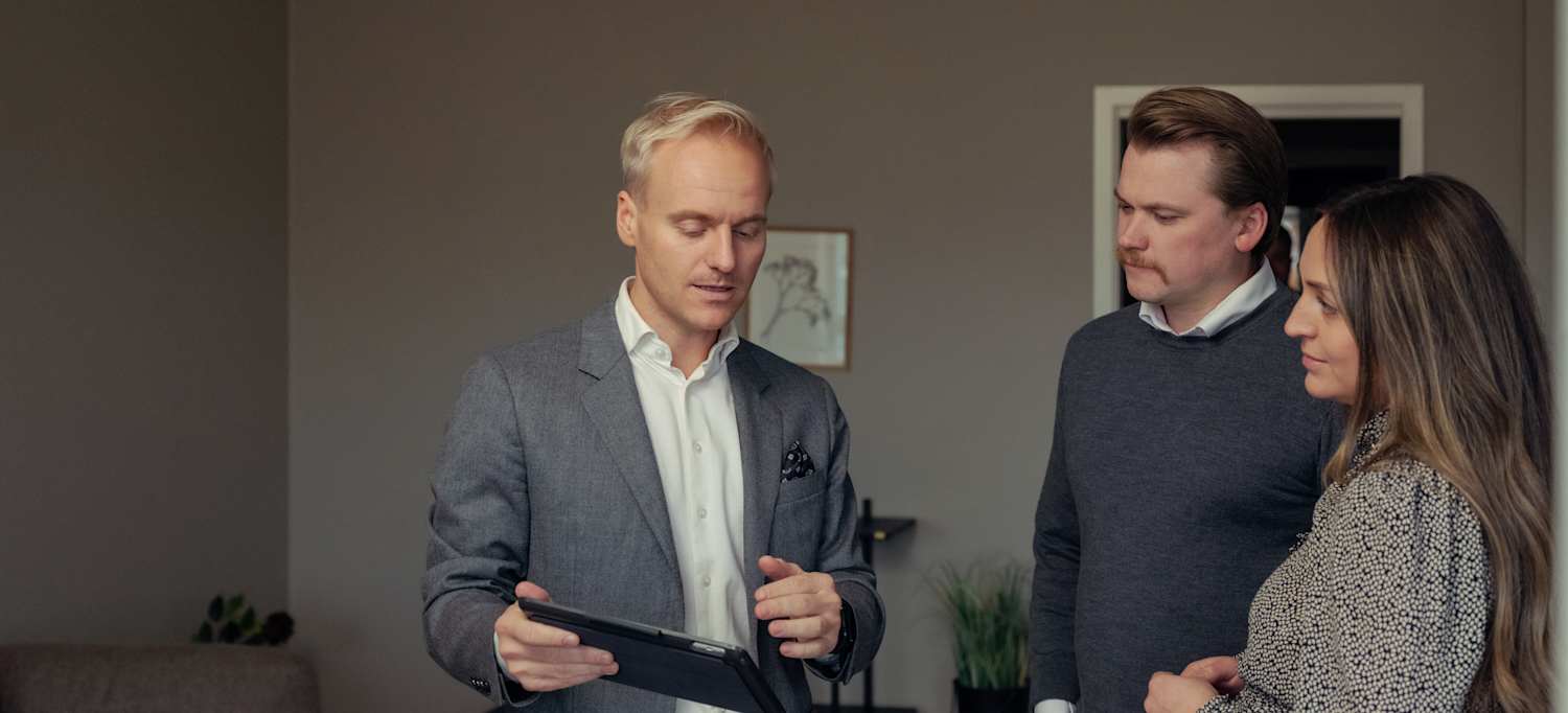 En eiendomsmegler står med en iPad som han viser fram til en dame og en mann mens han snakker. 