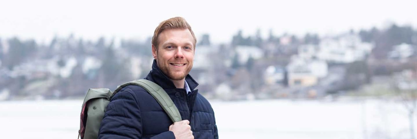 Mann med boblejakke og sekk smilende mot kamera. I bakgrunnen ser man et vann og et boligområde i det fjerne.
