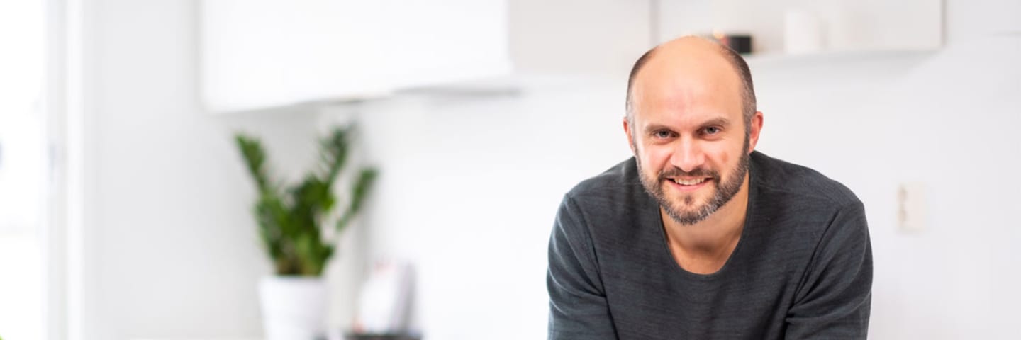 Fornøyd mann som lener seg smilende over en hvit kjøkkenbenk og ser inni kameraet.