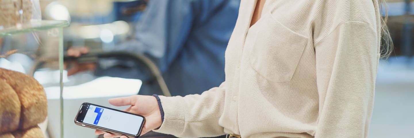 En fornøyd, blond kvinne står i kassen på et bakeri og gjør seg klar for kontaktløs mobilbetaling med Apple Pay