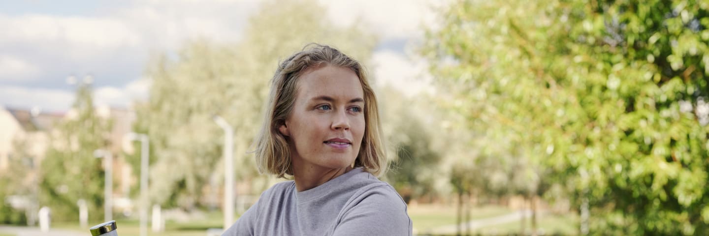 Ung kvinne holder en vannflaske i hånde og hviler seg mot rekkverket i en park.