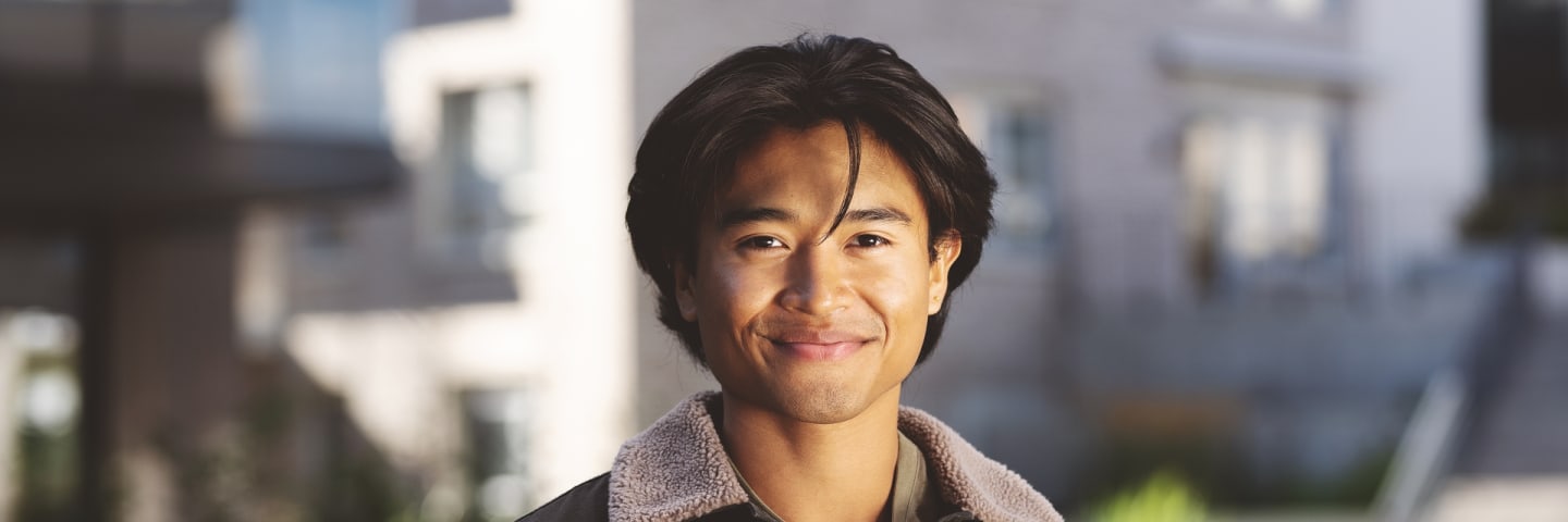 En ung voksen mann ser i kameraet og smiler. Han står ute og i front av en blokk.