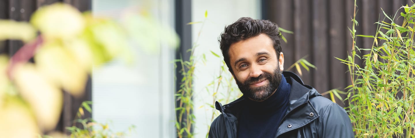 En mann ser mot kamera og smiler. Han står utenfor et hus. Det er grønne busker rundt og han holder en mobiltelefon i hånda.