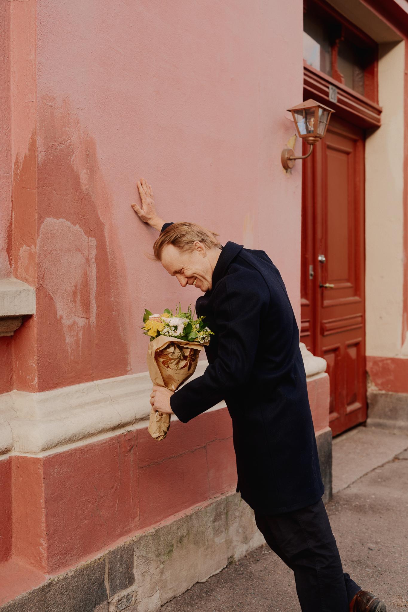 Thorbjørn Harr holder en blomsterbukett i hånda, han lukter på den mens han lener seg inn mot en rosa vegg.
