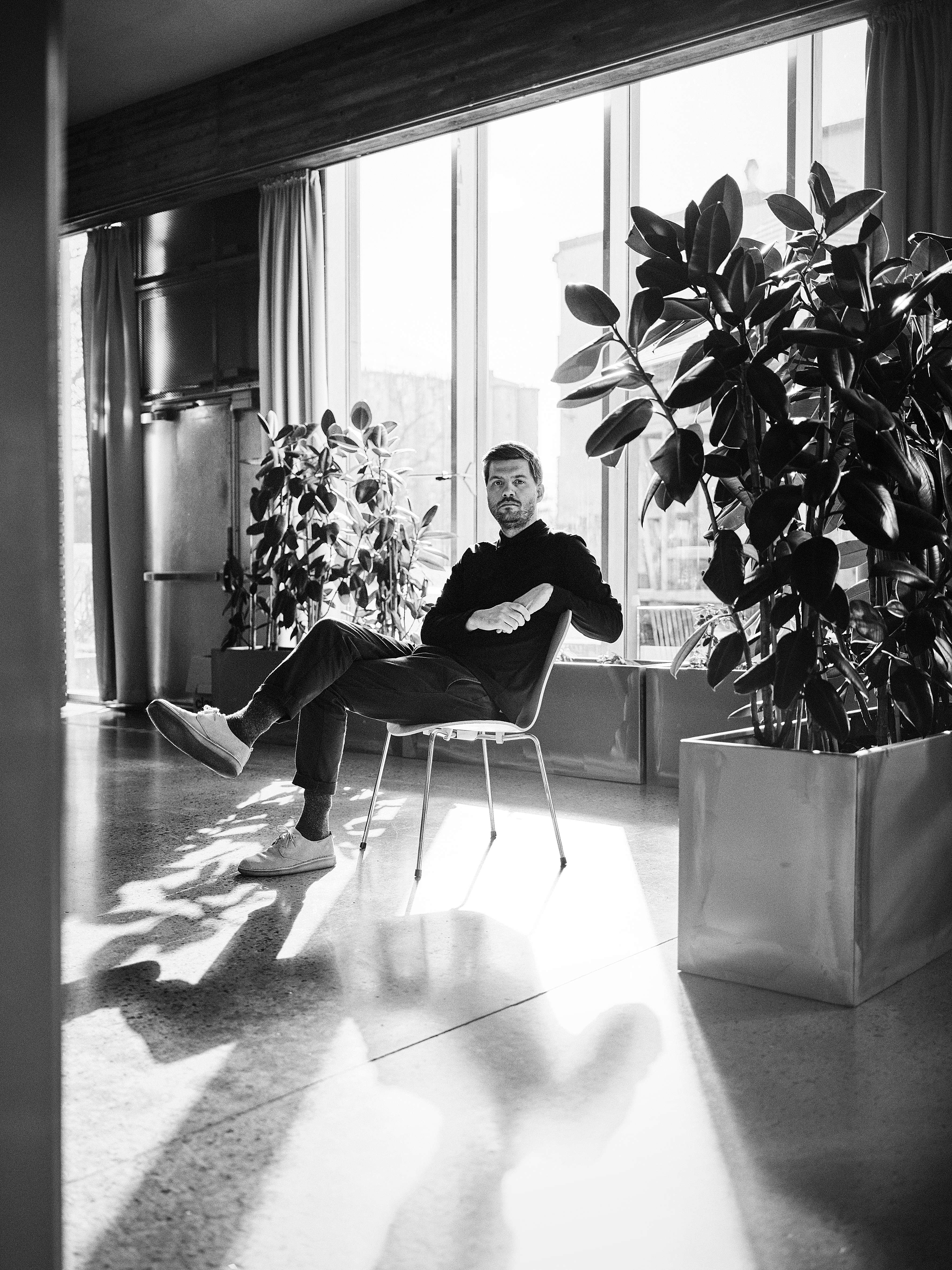 Et sort/hvitt bilde av en mann som sitter på en stol i et rom med flere planter.