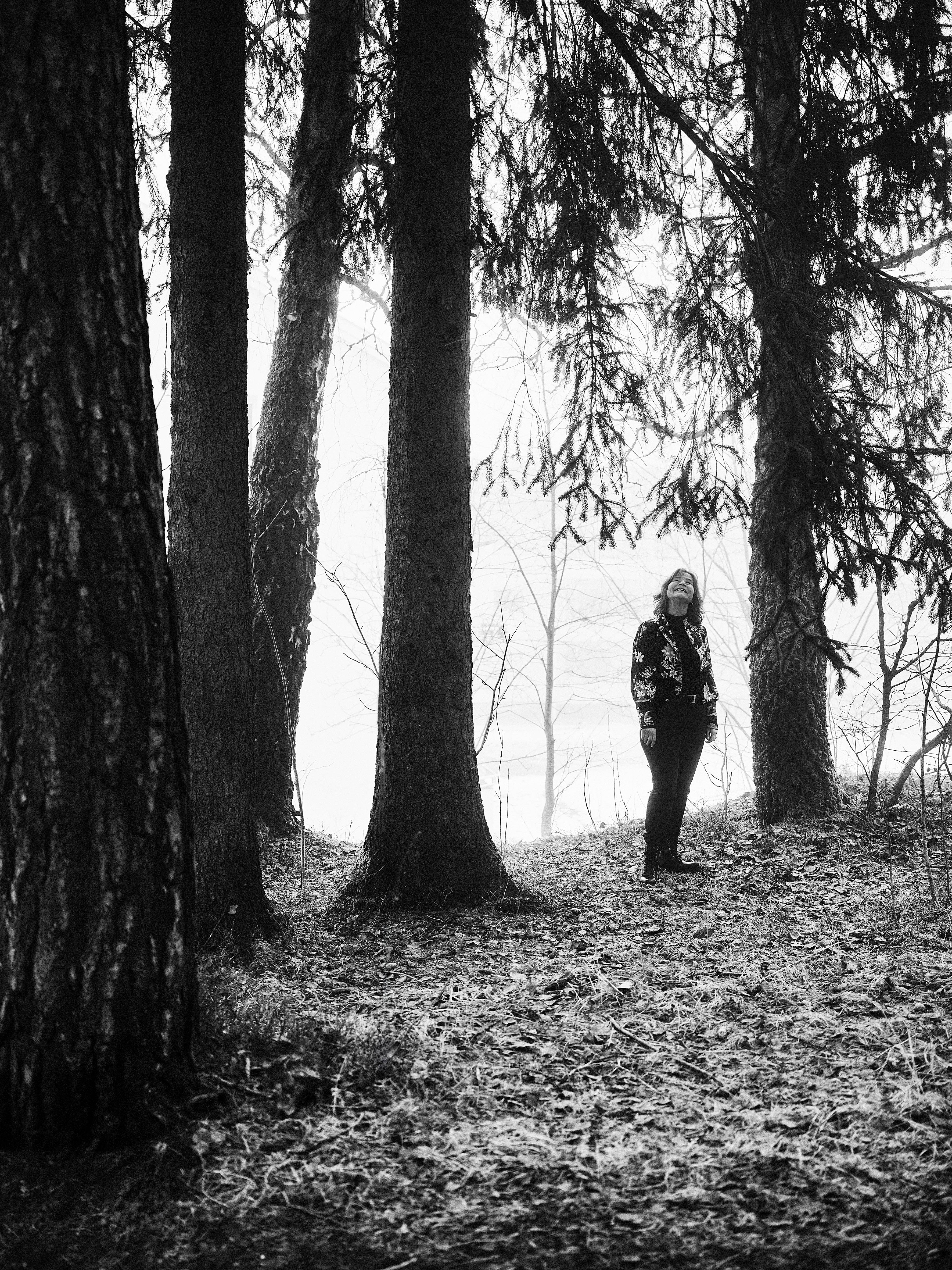 Et svart/hvitt bilde av en kvinne som står i en skog, på en bakke fylt med høstløv.