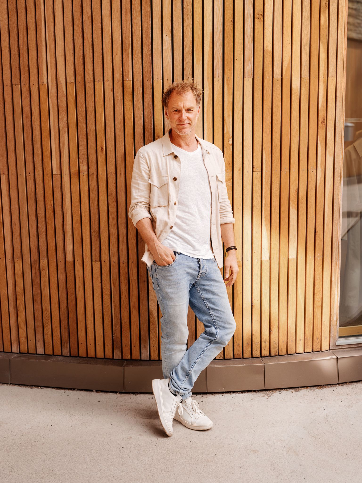 Hans Petter Aaserud har på seg en lys jeans, hvit t-skjorte og kremhvit overshirt. Han lener seg inntil en spilevegg.