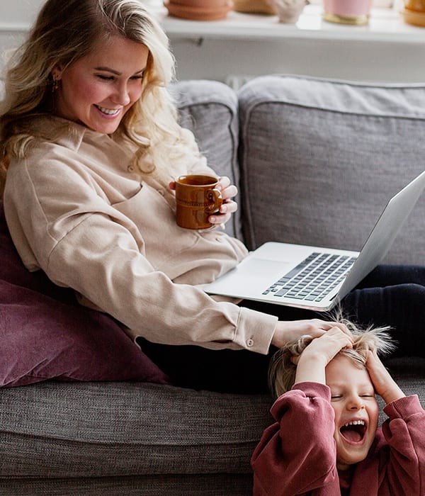 Mor sitter i sofaen med datamaskinen i fanget, mens sønnen leker og ler på gulvet.