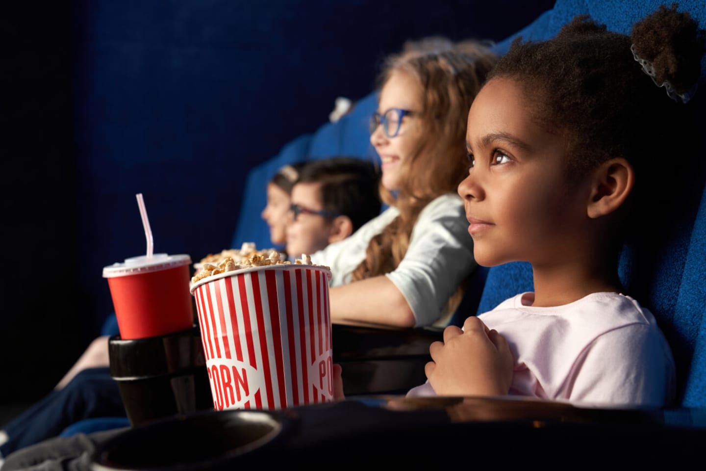 Tre barn sitter i en kinosal og ser på en film. De smiler. Hun ene jenta har popcorn og en annen jente har brus.