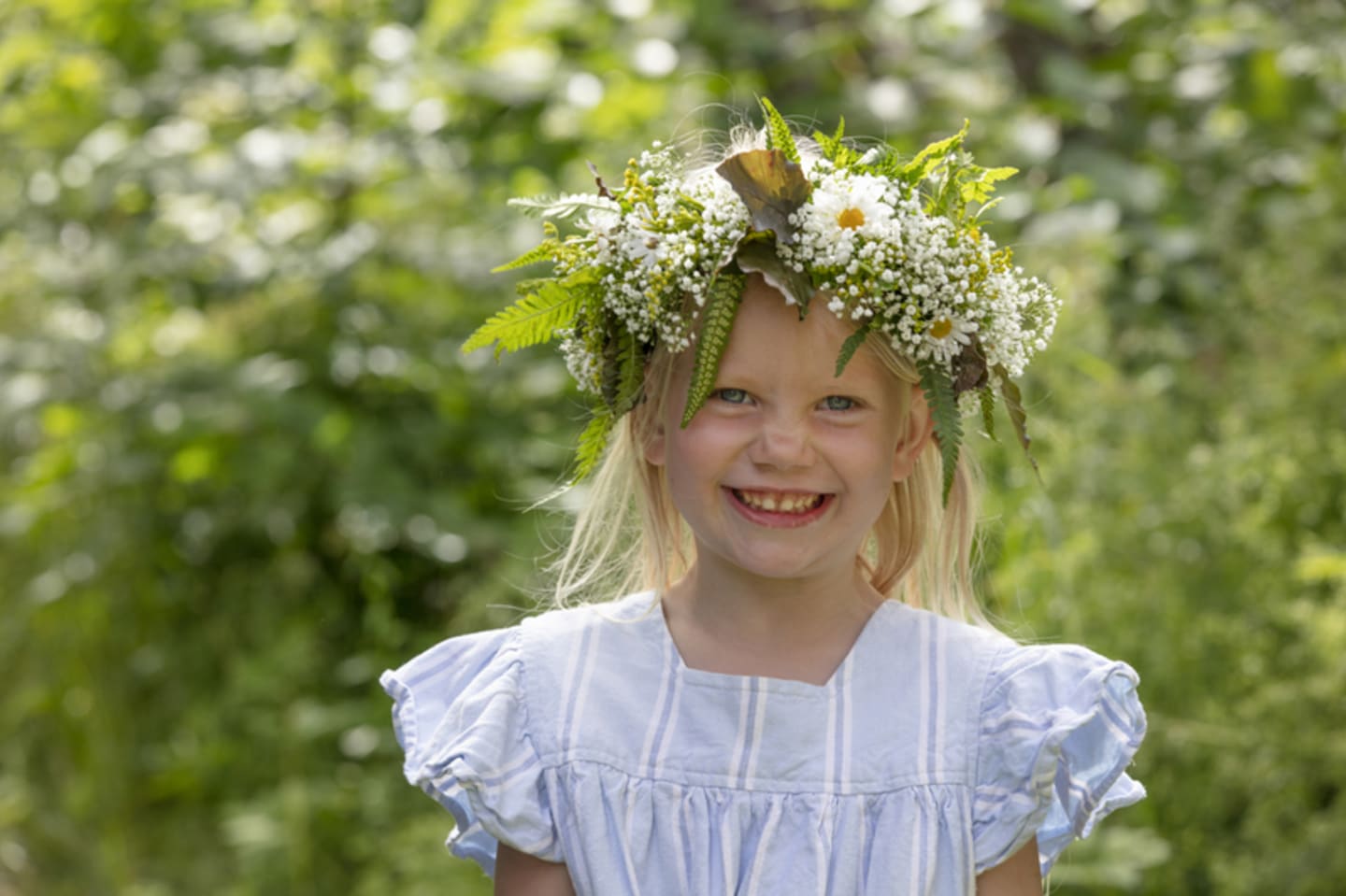 Foto av liten jente med blomsterkrans i en blomstereng som smiler til kameraet.