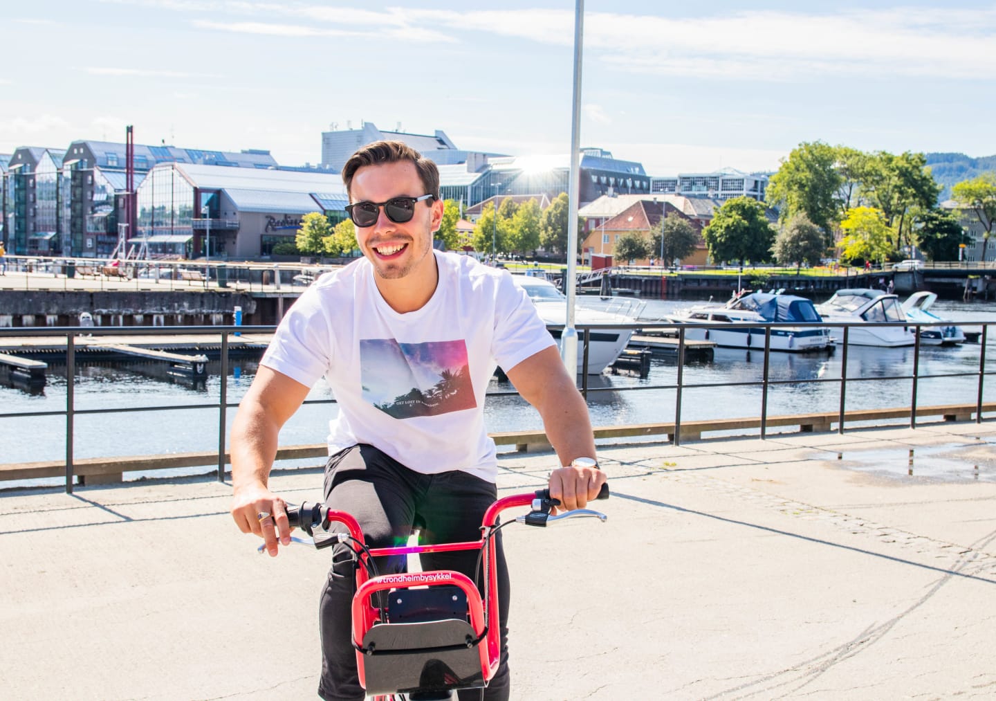 En ung mann sykler å en Trondheim Bysykkel. Det er fint vær, og han har på seg t-skjorte. Han smiler bredt og har på solbriller.