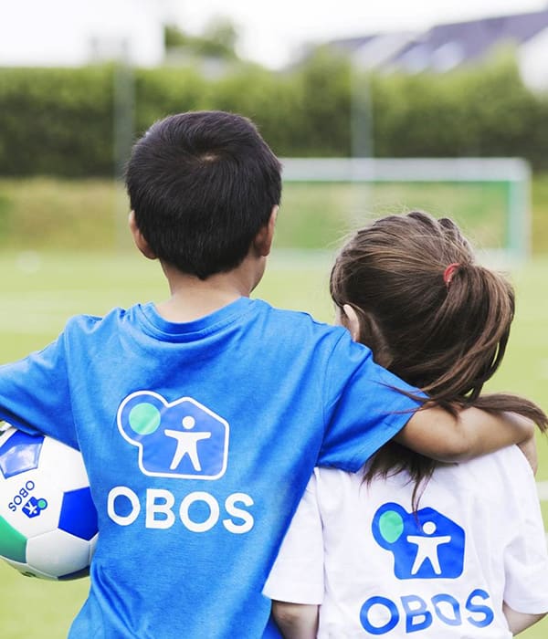 To barn med hver sin OBOS-trøye på seg som holder rundt hverandre mens de ser utover den grønne fotballbanen