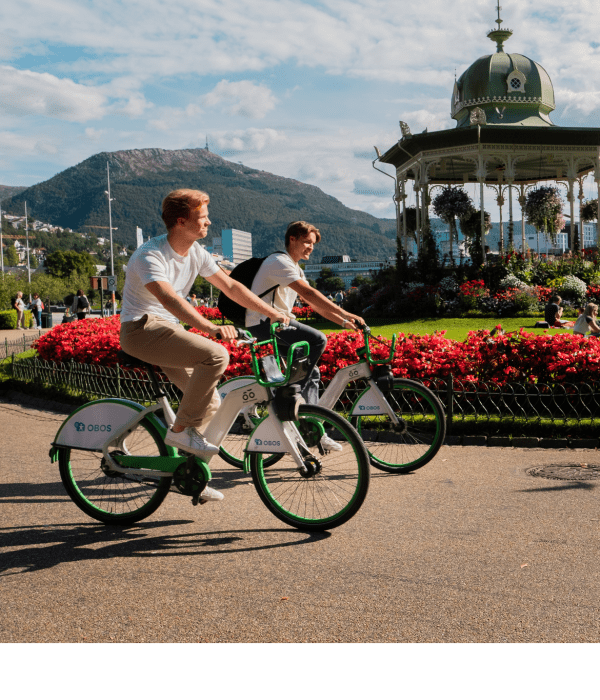 To gutter som sykler i solen midt i Bergen sentrum på hver sin hvite og grønne bysykkel.