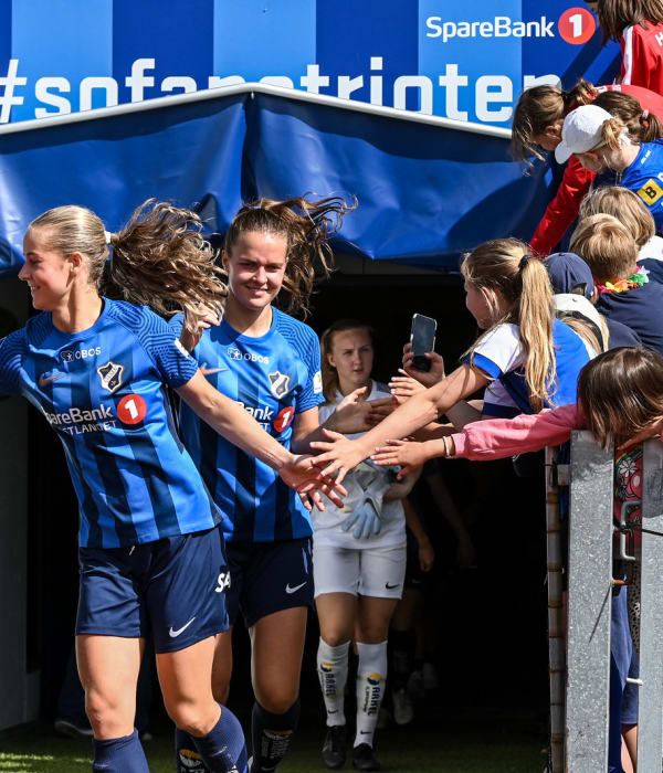 En gruppe jenter i stabæk fotballdrakt som løper på rekke ut på fotballbanen mens supportere tar hight five med dem.