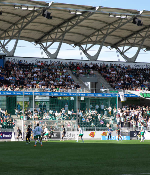 Bilde av HamKam supportere som står på tribunen rundt fotballbanen og heier på fotballspillerne på banen.