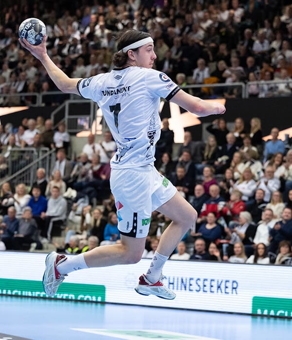 En håndballspiller hopper i det han skal kaste ballen mot mål. I bakgrunnen ser vi en lagkamerat og motstander som ser mot ham.