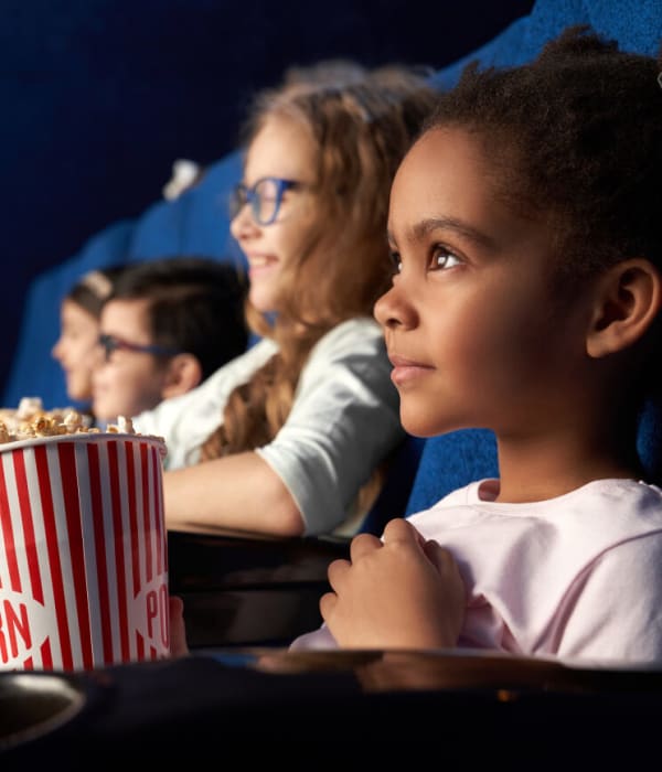 Tre barn sitter i en kinosal og ser på en film. De smiler. Hun ene jenta har popcorn og en annen jente har brus.