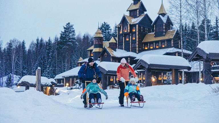 En familie står på kjelke. En mann og en dame har hvert sitt barn på hver sin kjelke og suser ut fra Hunderfossen. Du ser slottet i bakgrunnen.
