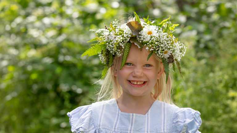 Foto av liten jente med blomsterkrans i en blomstereng som smiler til kameraet.