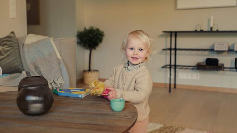En jente har på seg beige genser og rosa bukse. Hun smiler. Hun er i en leilighet.