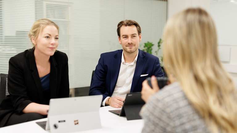 Tre personer sitter ved et bord. To personer sitter ved siden av hverandre og hjelper en over bordet. Han har på seg dress.