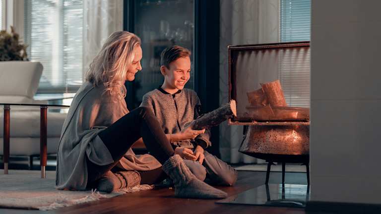 En dame og en gutt putter ved i en peis. Du ser lyset fra flammene. De smiler.