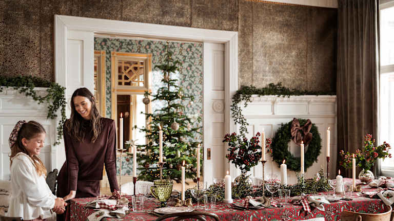 En kvinne og en jente står ved et fulldekket julebord i en stor stue. De er pyntet og smiler.