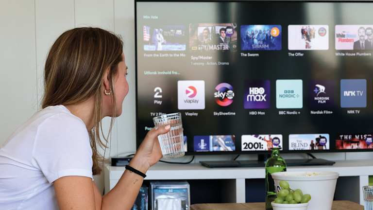 En kvinne ser på TV. Hun har strømmetjeneste. Hun holder et glass vann.
