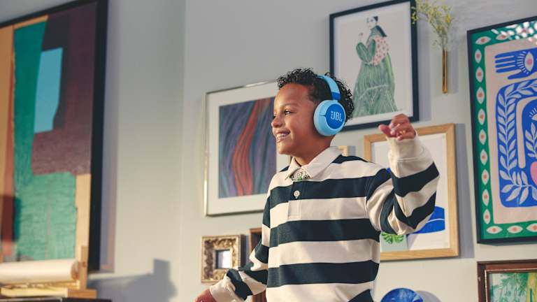 Et barn smiler bredt og danser litt med JBL hodetelefoner på hodet. Det er masse kunstverk på veggene bak han.