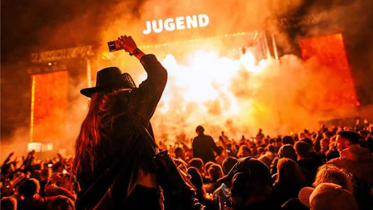 Festivalscenen på Jugend. Det er flammer fra scenen og en person sitter på skuldrene til en annen og holder hånden opp i været.
