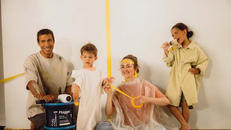 Fire mennesker har kledd seg for å male. Noen har teip i ansiktet og på veggen. De har malingsspann og pensler, og smiler.