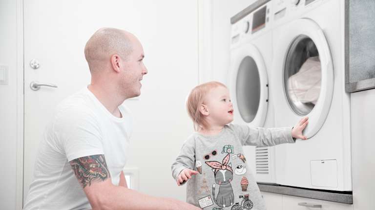 Far og barn vasker klær. Barnet ser på klærne inni vaskemaskinen.