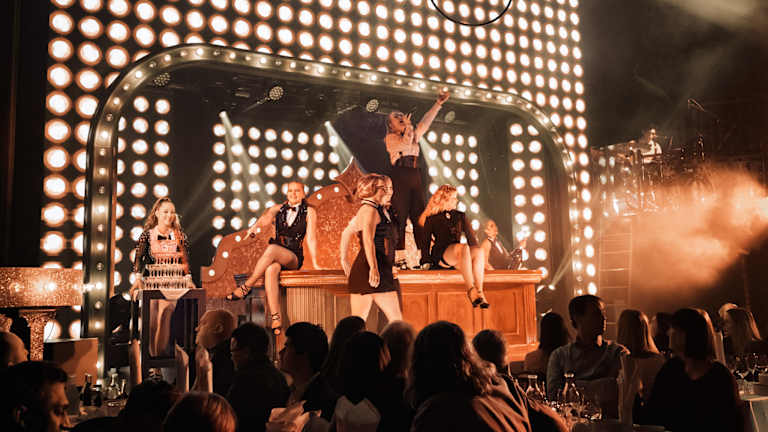 Du ser oppimot syv personer som opptrer på en scene. Damene har på seg festklær. Rundt dem sitter det folk rundt bord og ser på.