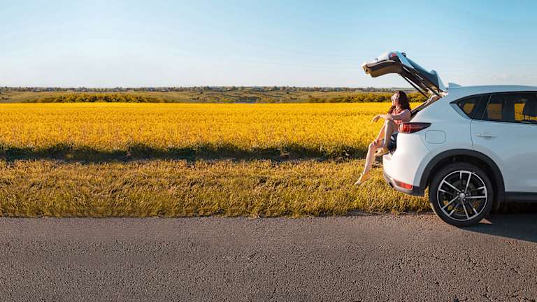 En dame sitter i bagasjerommet på en hvit bil og titter utover landskapet. Hun har på seg sommerklær og solbriller.