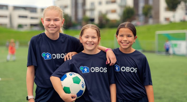 Tre fotballjenter står på en fotballbane. De ser i kameraet og smiler. Foto.