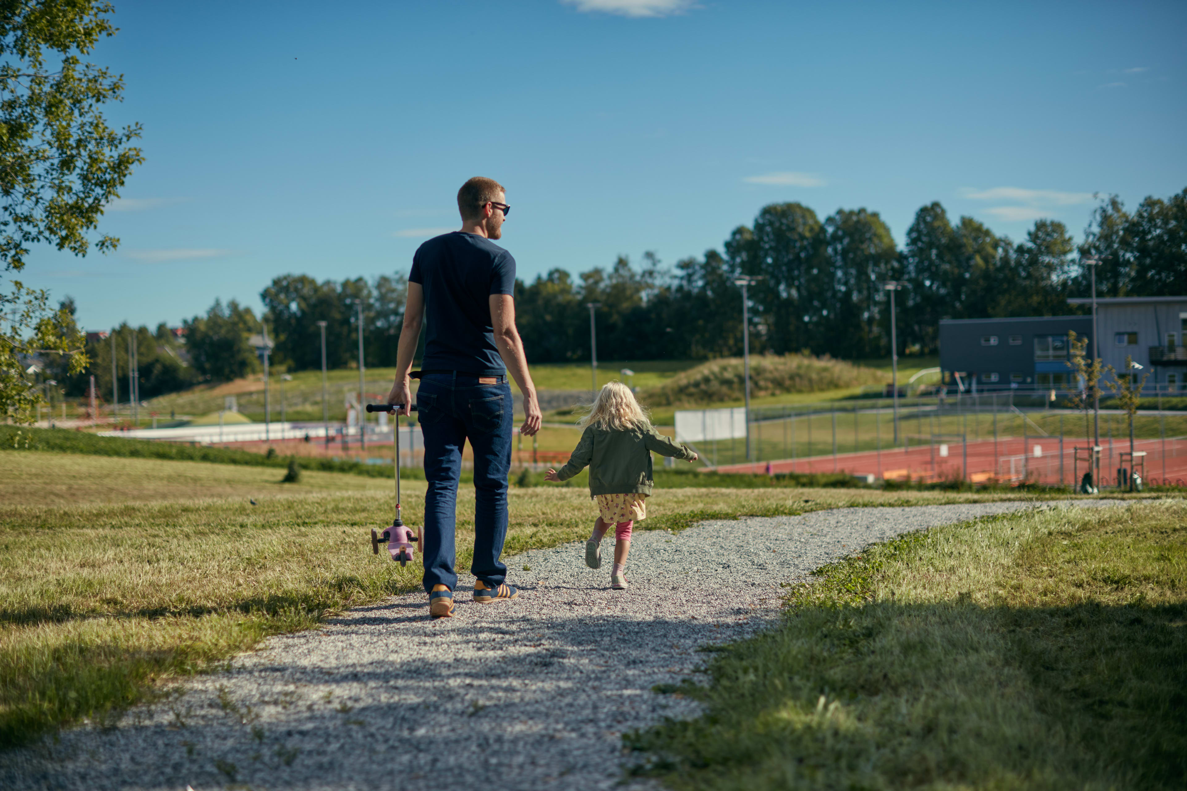 En far som bærer på sparkesykkel, mens datteren løper mot lekeplassen.