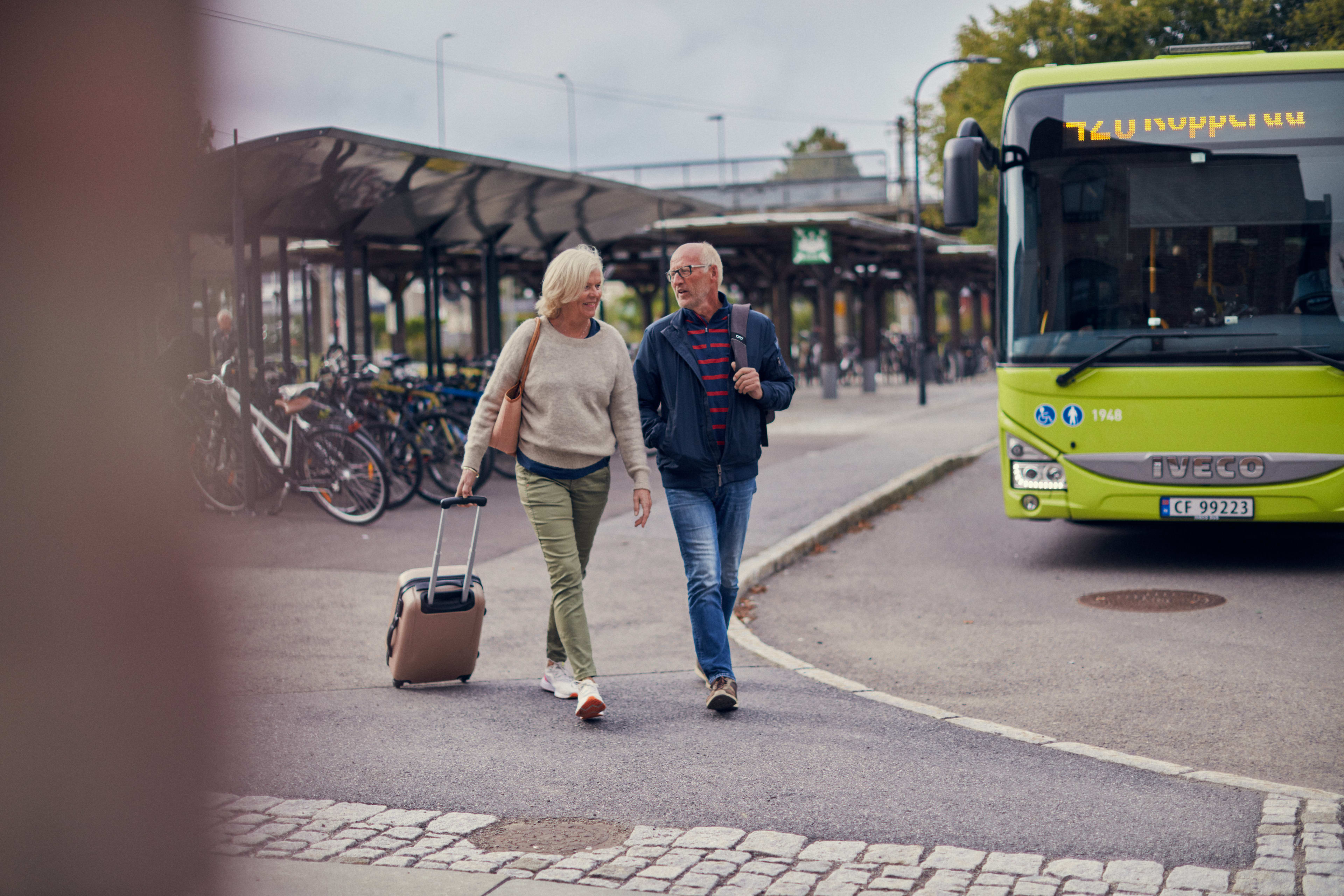 Et par som går med koffert på bussholdeplassen på Jessheim.