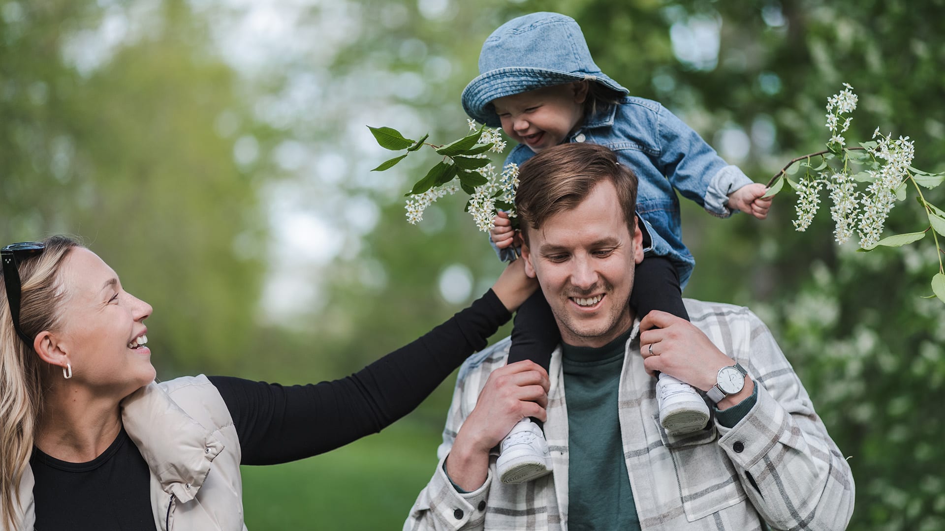 Foto på en familj med en pappa som bär ett barn på sina axlar och en mamma som håller i barnets ben, alla skrattar.